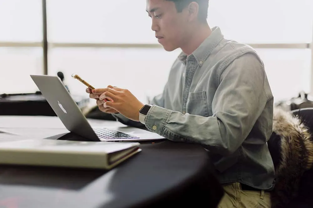 Man sitting at desk with a laptop and checking his mobile phone